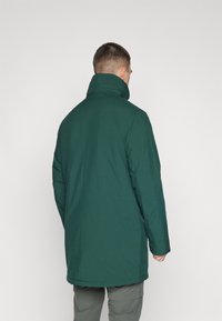 Grønn vanntett parka med høy krage, med en glatt tekstur, rett snitt og sidelommer. Designet er enkelt og funksjonelt.