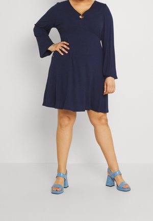Femme portant une robe bleu marine à manches longues avec un décolleté en encoches et des sandales à talons hauts bleu ouvertes à l'avant sur un fond uni.
