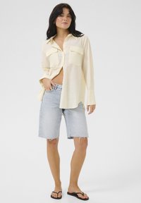 Camicia gialla chiara a maniche lunghe con bottoni, dotata di due tasche frontali, abbinata a pantaloni corti in denim azzurro e ciabatte nere. Vestibilità casual.