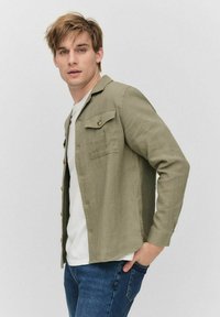 Olijfgroen linnen shirt met twee borstzakken, knoopsluiting aan de voorkant en lange mouwen, gedragen over een wit T-shirt en blauwe jeans.