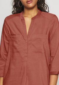 Blouse couleur rouille en tissu léger, avec un décolleté en V, deux poches poitrine et des manches ¾. Texture lisse avec un léger éclat.