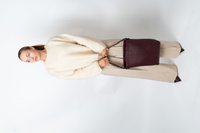 Mulher com suéter creme e calças largas bejes, segurando uma grande bolsa de couro marrom escuro, em pé contra um fundo branco.