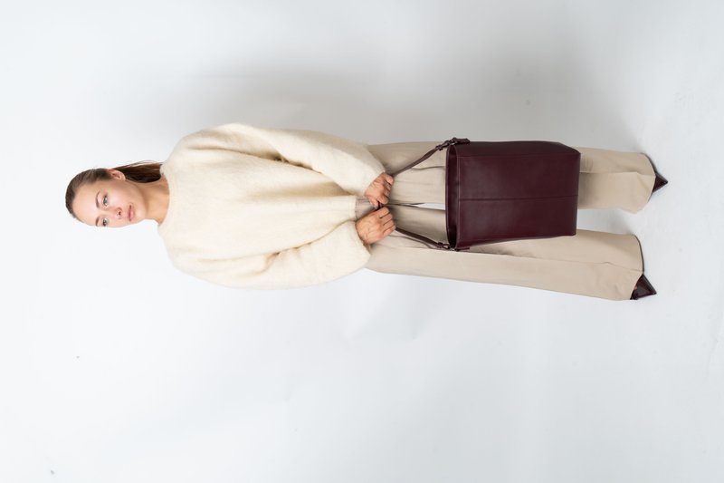 Mulher com suéter creme e calças largas bejes, segurando uma grande bolsa de couro marrom escuro, em pé contra um fundo branco.