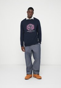 Granatowy sweatshirt z różowym graficznym wzorem drzewa i napisem "TIMBERLAND", połączony z niebieskimi dżinsami i beżowymi butami. Gładka faktura materiału.