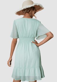 Vestido verde menta con mangas cortas de volantes, cintura fruncida y dobladillo con volantitos. La tela ligera añade una textura fluida.