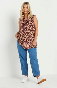 Mouwloze blouse in burgundy en beige abstract patroon, gecombineerd met lichtblauwe jeans. Model staat met handen in de zakken, draagt witte sneakers.