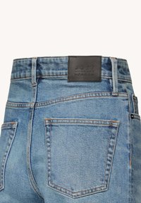 Vaqueros de mezclilla azul con corte ajustado, cuentan con un parche de cuero negro con texto en relieve, herrajes metálicos y costuras detalladas en los bolsillos.