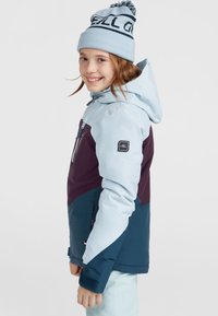 Veste de ski bleue et violette avec capuche, dotée de poches zippées et d'un écusson, associée à un bonnet en tricot bleu clair avec un pompon.