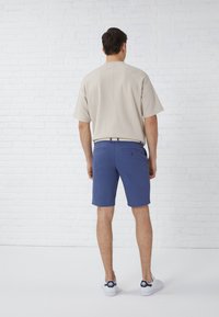 Beige Kurzarmhemd, lockere Passform; blaue Shorts, gerader Schnitt; weiße Sneaker mit marineblauen Akzenten; strukturierter Gürtel.