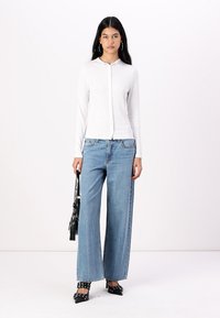 Haut blanc à manches longues et boutonné avec un tissu texturé ; associé à un jean large bleu clair. Chaussures noires à bout pointu avec des accents cloutés.