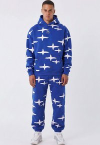 Sudadera con capucha azul y pantalones a juego con un patrón de estrellas blancas. El material parece suave, con un ajuste holgado y puños elásticos. Bolsillo frontal en la sudadera.