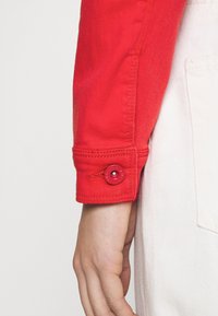 Chaqueta de mezclilla roja con detalle de puños abotonados, que presenta un botón rojo redondo con superficie texturizada y una prenda blanca contrastante debajo.