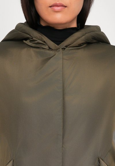 Frau in einem olivgrünen Kapuzenmantel über einem schwarzen Rollkragenpullover, mit geschlossenen Lippen und frontal gewandt.