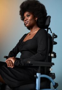 Αυτοπεποίθηση γυναίκα με φυσικά μαλλιά, φορώντας ένα μαύρο φόρεμα, καθιστή με ευθύ τρόπο σε ένα σύγχρονο αμαξίδιο, με φόντο σε γραδίαντ.