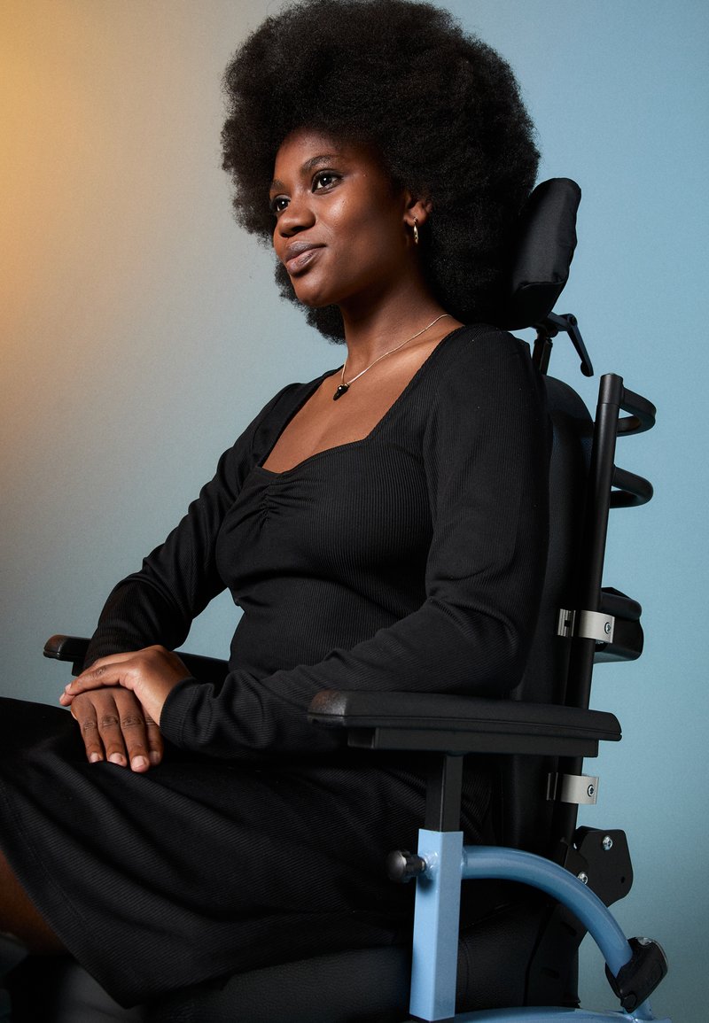 Αυτοπεποίθηση γυναίκα με φυσικά μαλλιά, φορώντας ένα μαύρο φόρεμα, καθιστή με ευθύ τρόπο σε ένα σύγχρονο αμαξίδιο, με φόντο σε γραδίαντ.