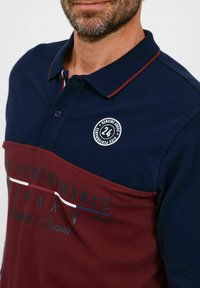 Polo majica u tamnoplavoj i burgundskoj boji, s logotipom, dugmadi i tekstualnim detaljima u kontrastnim bojama. Dizajn s kratkim rukavima.
