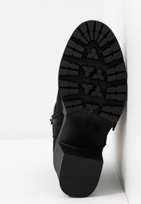 Semelle de bottine noire avec un talon épais et un motif de bande de roulement profond et multidirectionnel, appuyée en angle contre un mur blanc.