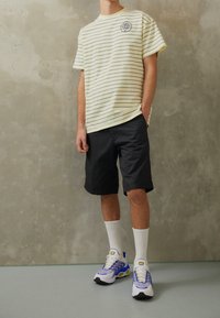 Randig gul t-shirt med logotyp på bröstet, svarta shorts, vita strumpor och flerfärgade träningsskor. Har en avslappnad passform och en avslappnad stil.