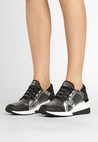 Piernas con zapatillas bajas negras y plateadas con suelas blancas, de pie sobre una superficie gris contra un fondo blanco.
