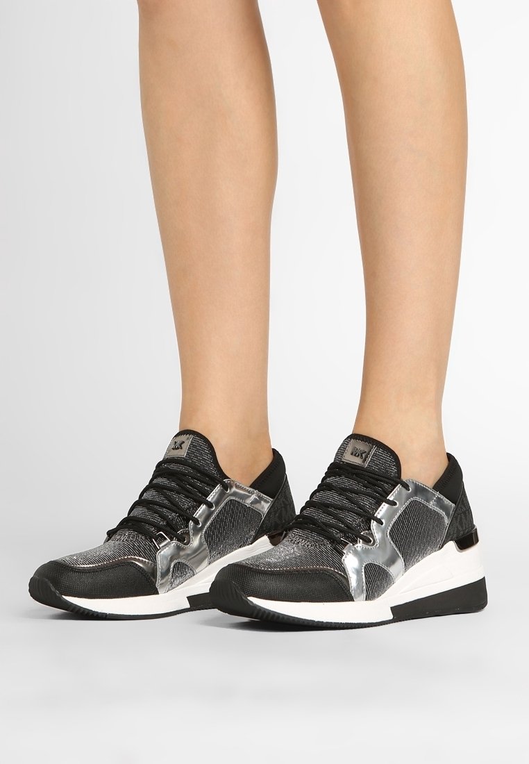 Piernas con zapatillas bajas negras y plateadas con suelas blancas, de pie sobre una superficie gris contra un fondo blanco.