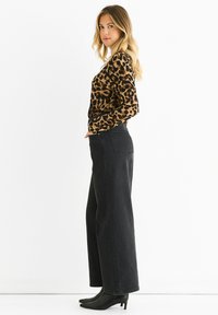 Top a maniche lunghe con stampa leopardo abbinato a jeans neri a vita alta e gamba larga. Il modello indossa stivaletti neri con tacco, in piedi contro uno sfondo bianco.