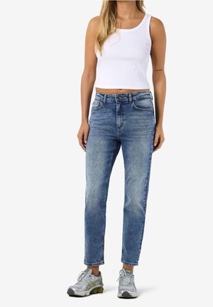 Femme portant un crop top blanc sans manches, un jean bleu taille haute et des chaussures de sport argentées, debout devant un fond uni.
