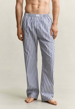 Man draagt losse blauw-wit verticale gestreepte pyjamabroek, staat blootsvoets op een effen achtergrond.