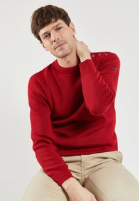 Roter Strickpullover mit Rundhalsausschnitt, gerippten Bündchen und zwei Knopfakzenten an der linken Schulter. Kombiniert mit beigefarbenen Hosen, glatte Textur.