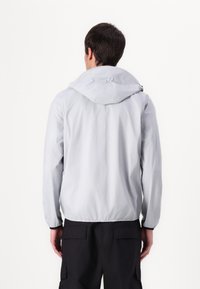 Jachetă impermeabilă de culoare gri deschis, cu glugă ajustată, mansete elastice și margine strânsă. Textură netedă, design minimalist și fără brand vizibil.