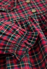 Tissu à carreaux rouge, vert et bleu marine avec poignet froncé et boutons en bois, montrant les coutures détaillées et la texture du vêtement.