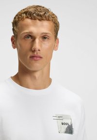 Hvid bomulds t-shirt med rund hals. Har et grafisk print på venstre bryst, inklusive ordet "BOSS" og abstrakte designelementer.