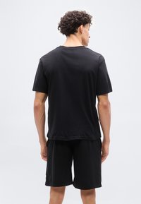 Άνδρας με σγουρά μαλλιά, φορώντας μαύρο μπλουζάκι και μαύρο σορτς, στέκεται αντιμετωπίζοντας ένα απλό ανοιχτό γκρι φόντο.