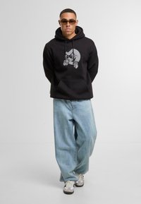 Felpa nera con una grafica di un personaggio in grigio, abbinata a jeans baggy azzurri chiari e sneakers beige. Gli accessori includono occhiali da sole.