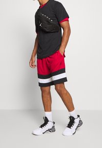 Man som bär en svart och röd lagertröja, röda basketshorts med svarta och vita ränder, vita högstövlade sneakers och en svart axelremsväska.