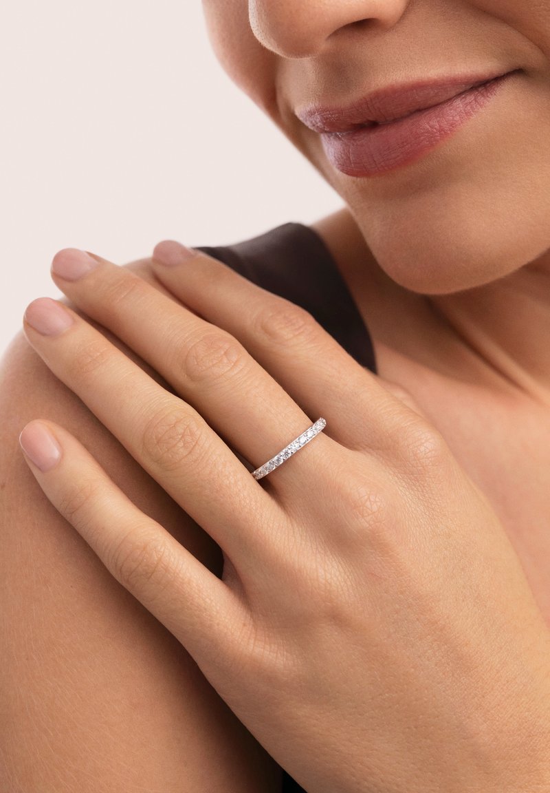 Anillo de plata con una sola fila de piedras preciosas redondas y transparentes, llevado en una mano con uñas cortas y naturales, sobre un fondo neutro.