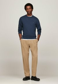 Maglione blu navy a maglia con polsini e orli a coste, abbinato a pantaloni beige plissettati e stivali neri con suola spessa. Camicia bianca semplice sotto.