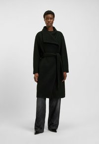 Czarny, teksturowany płaszcz zapinany z szerokim kołnierzem i dopasowanym paskiem; modelka nosi go z czarnymi szerokimi spodniami i butami z wydłużonym noskiem.