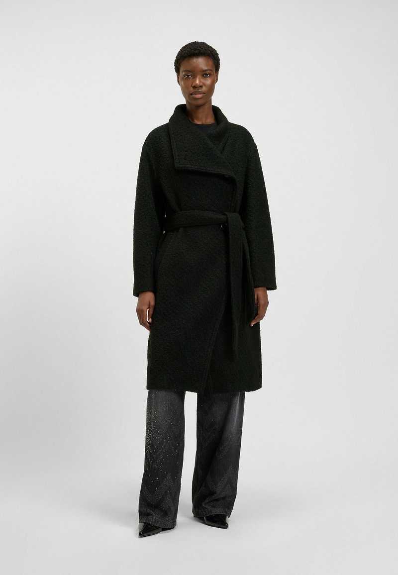 Czarny, teksturowany płaszcz zapinany z szerokim kołnierzem i dopasowanym paskiem; modelka nosi go z czarnymi szerokimi spodniami i butami z wydłużonym noskiem.