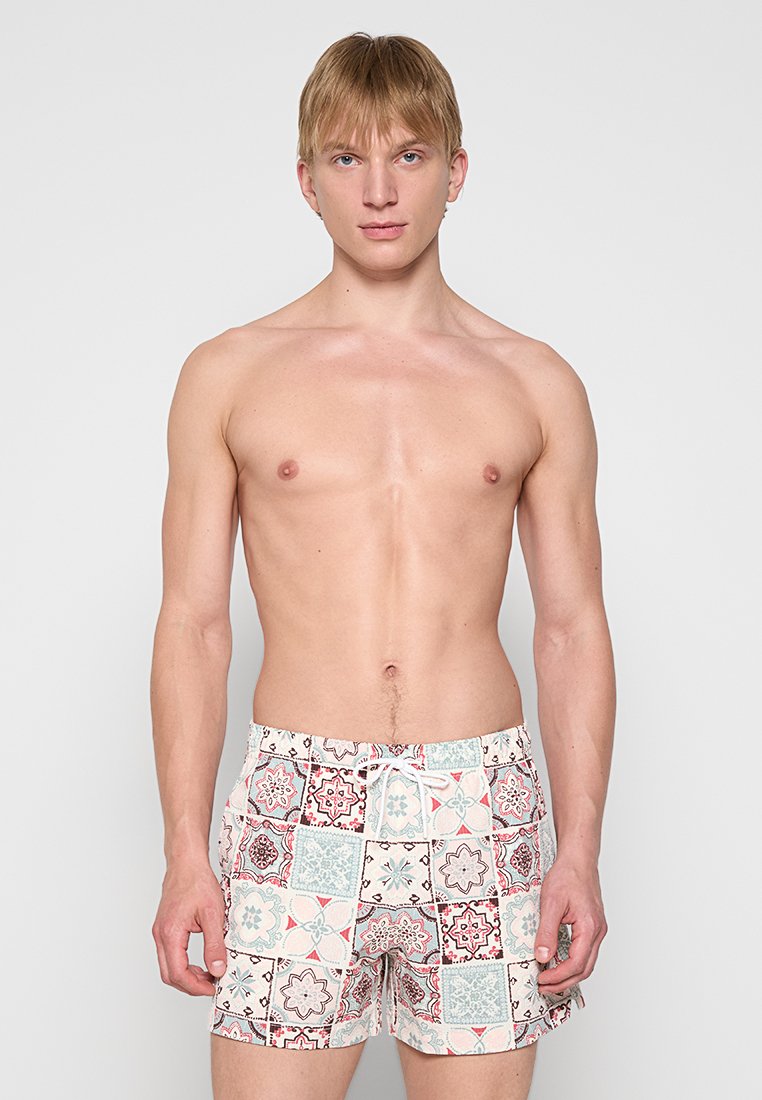 Un homme blond se tenant contre un fond uni, portant un short de bain à motifs avec des designs en carreaux géométriques dans des tons rouge, bleu et beige.