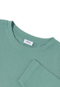 Chemise à manches longues verte à côtes avec un col rond et une poche plaquée. L'étiquette indique "CECIL" sur une étiquette rectangulaire blanche.