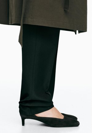 Jambes inférieures portant un pantalon noir ajusté et des chaussures en daim noires à talons bas, avec un ourlet de manteau olive foncé visible au-dessus.