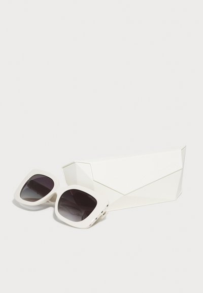 Lunettes de soleil géométriques blanches surdimensionnées avec des verres foncés reposant dans un étui rigide angulaire blanc assorti sur un fond uni.