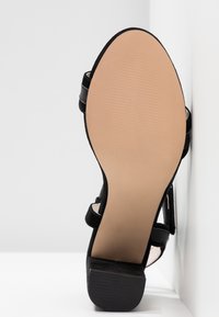 Vue de la semelle d'une sandale noire à talon avec une semelle extérieure lisse beige et un talon bloc, positionnée à la verticale sur un fond d'angle blanc.