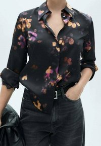 Mujer vistiendo una blusa negra de botones con patrones florales morados y naranjas, mangas remangadas, metida en jeans negros con un cinturón.
