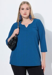 Top de patrón espigado azul con mangas tres cuartos, escote en V y un corte holgado, combinado con un pequeño bolso negro.