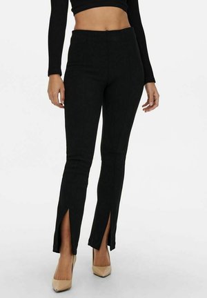 Mujer vistiendo pantalones negros de cintura alta con aberturas frontales cerca de los tobillos, combinados con tacones beige de punta puntiaguda y un top corto negro.