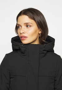 Chaqueta aislante negra con un alto cuello y capucha ajustable, que cuenta con dos bolsillos frontales y una textura de tela suave.