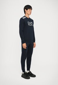 Felpa navy con joggers coordinati, in misto cotone, polsini a coste, con logo sul petto e dettagli a strisce sulle spalle, abbinata a sneaker nere.