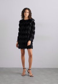 Μαύρο φόρεμα από ψεύτικη γούνα με ζιγκ-ζαγκ υφή, μακριά μανίκια και χαλαρή εφαρμογή, συνδυασμένο με μαύρες ψηλές γόβες με λουράκια. Ουδέτερο φόντο.