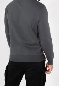 Sudadera gris con un cuello y puños acanalados, con una etiqueta en el lateral. Combinada con pantalones cargo negros. Textura suave, corte casual.